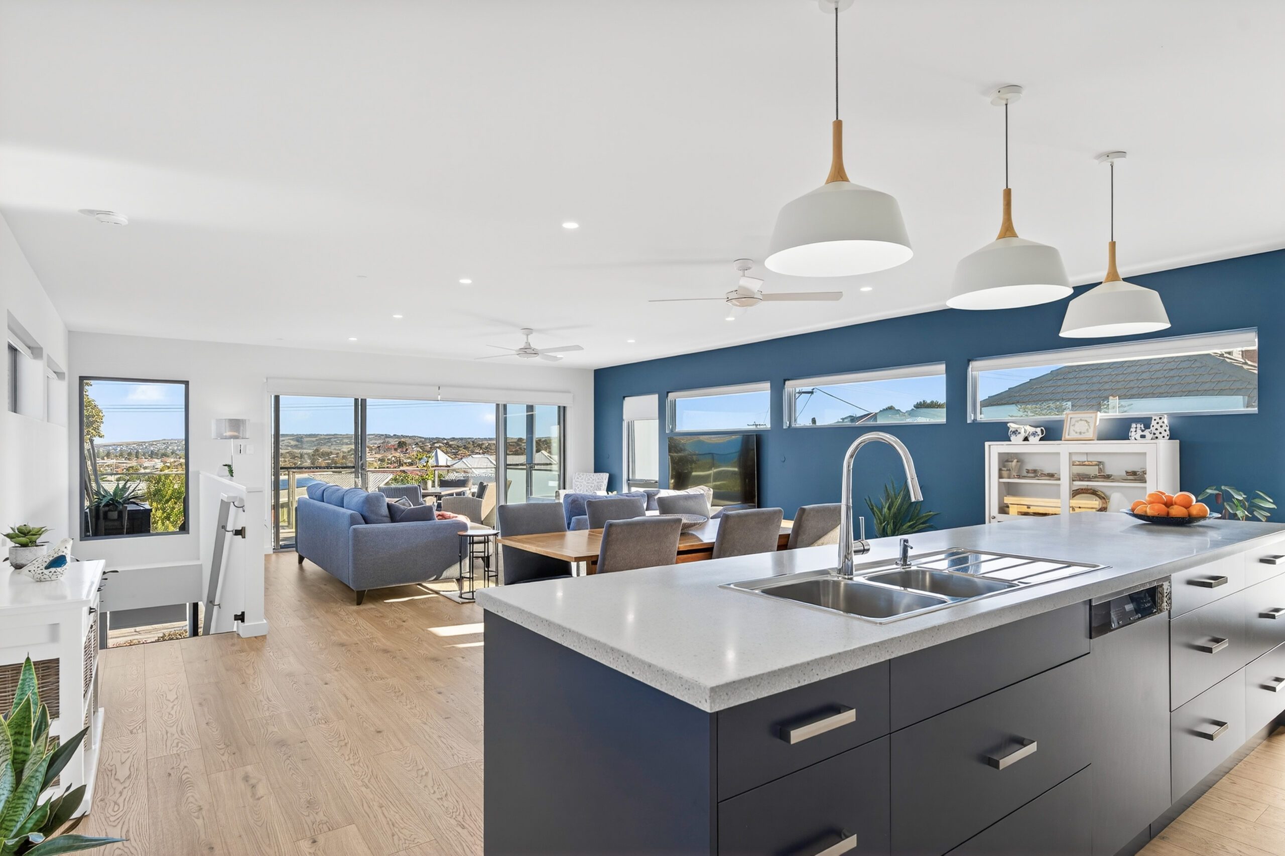 Open-plan kitchen and living area at Cudmore Rd, McCracken, with a navy feature wall, stone island bench, timber pendant lights and coastal views through wide windows.