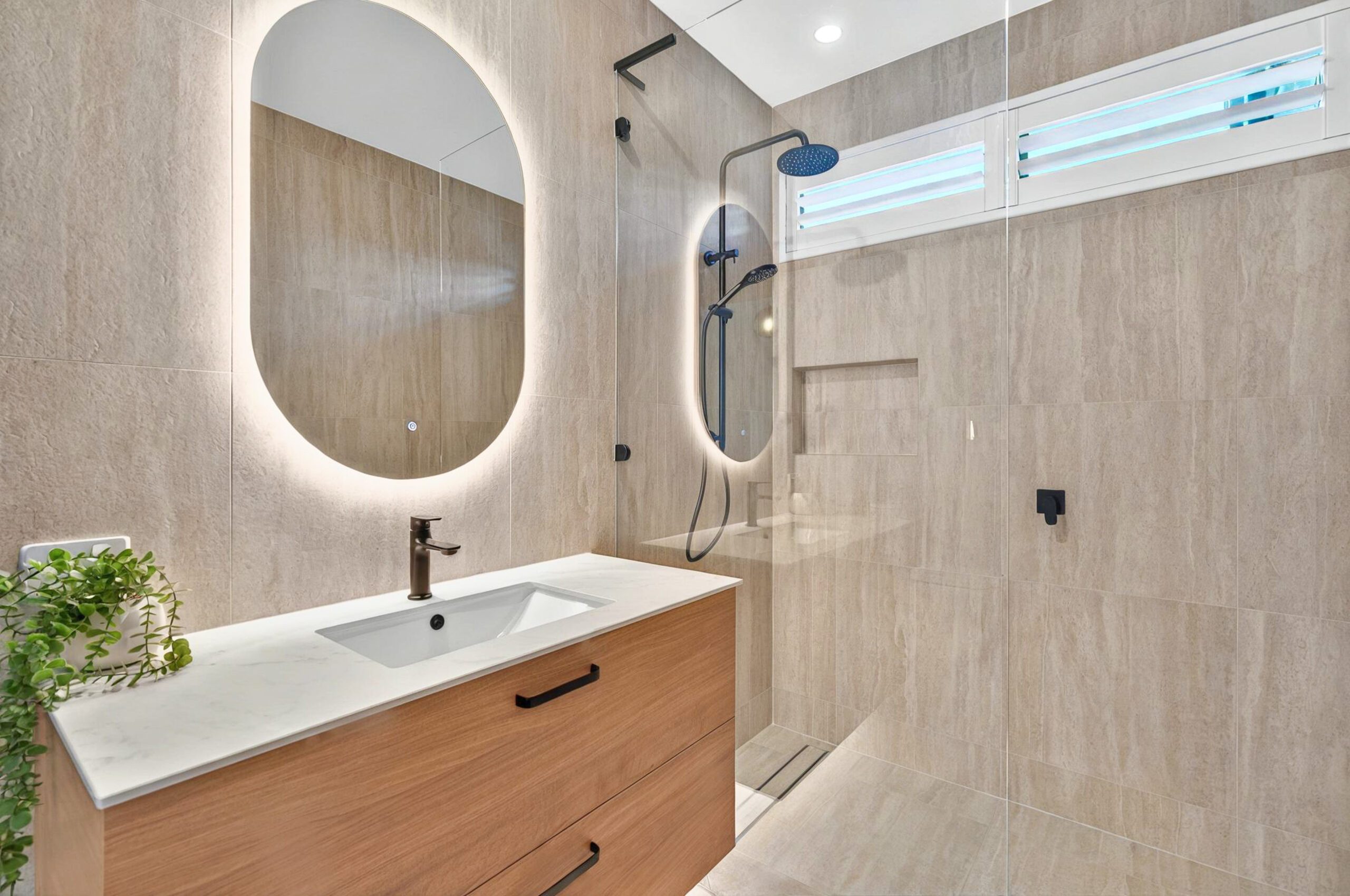 Ensuite at Musk Duck Ave, Chiton, with a backlit oval mirror, timber vanity, stone-look tiles, black tapware and a walk-in shower.