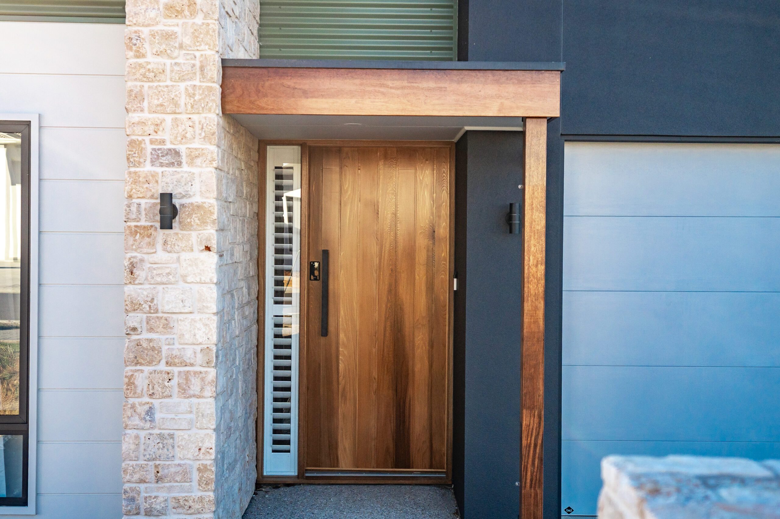 Front entry at Musk Duck Ave, Chiton, with a timber pivot door framed by a natural stone pillar, weatherboard cladding and dark render.