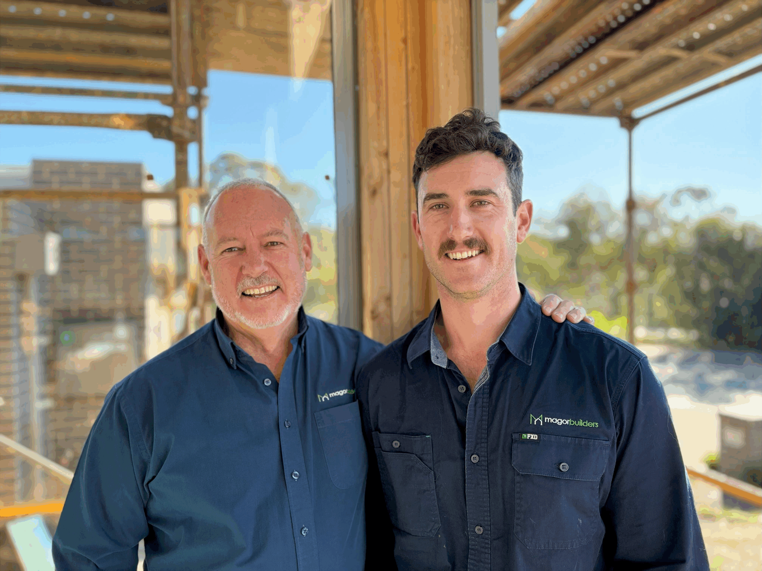 Simon and Liam Magor of Magor Builders standing together on a timber-framed building site, both wearing navy Magor Builders work shirts.